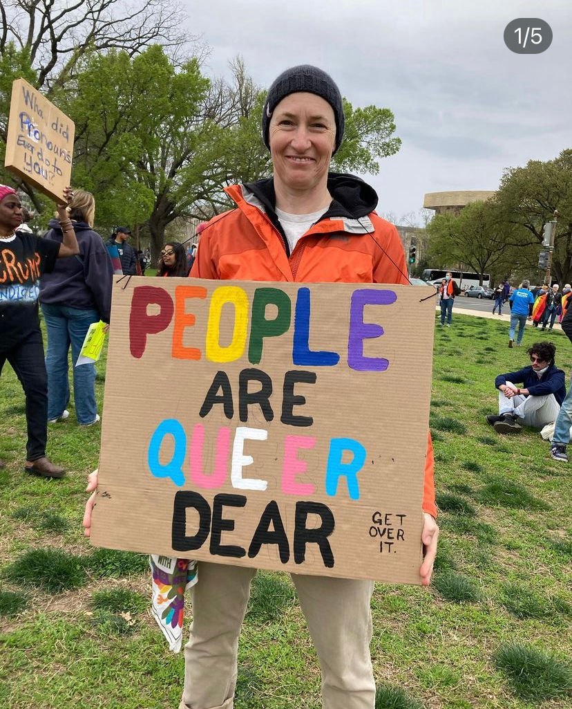 the author stands with a background of a dwindling crowd of protestors. they are smiling and holding a sign that says "People are queer dear."