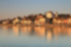 Sunlit town by a lake with historic buildings and a castle, reflecting warm tones in the water during a peaceful golden-hour scene.