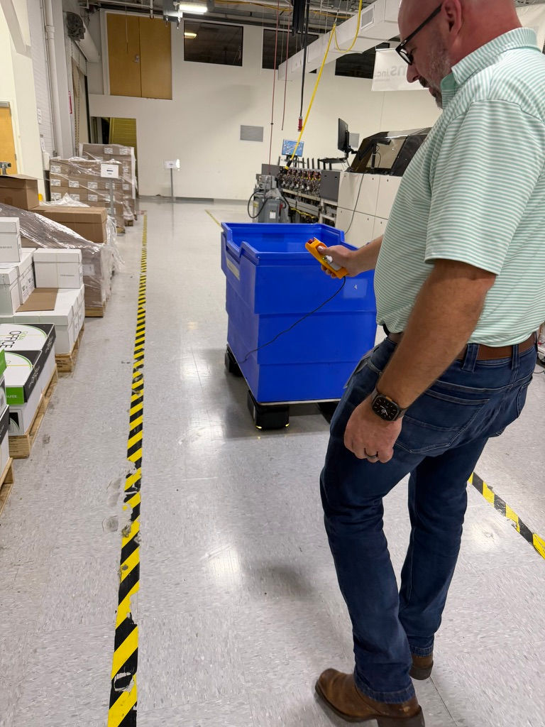 Man in a warehouse using a handheld device to scan a blue bin on wheels. Boxes are stacked on pallets. Yellow-black tape marks floor.
