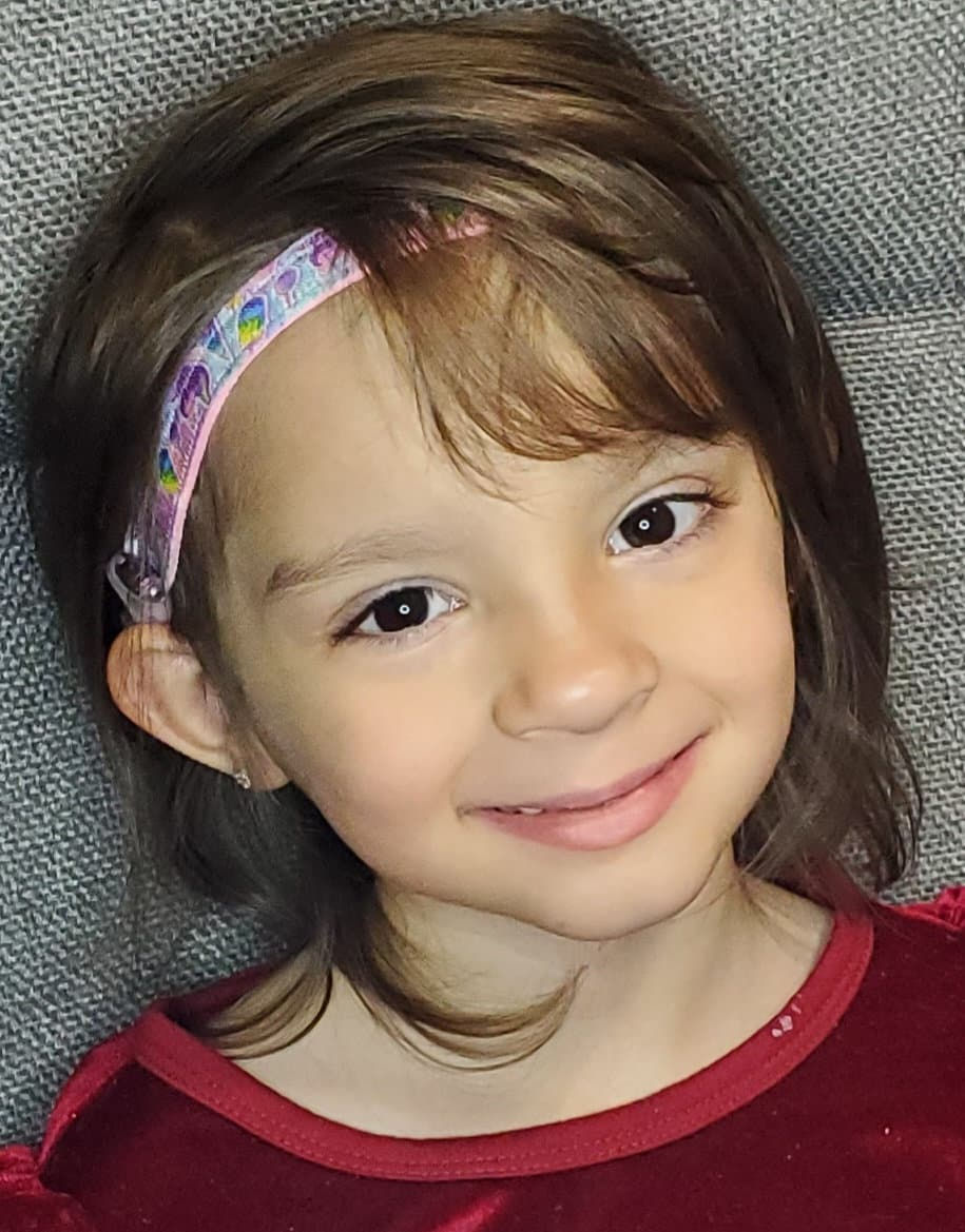Young girl smiling with a hearing aid and a patterned headband. Snowman Hearing Aid Charms.