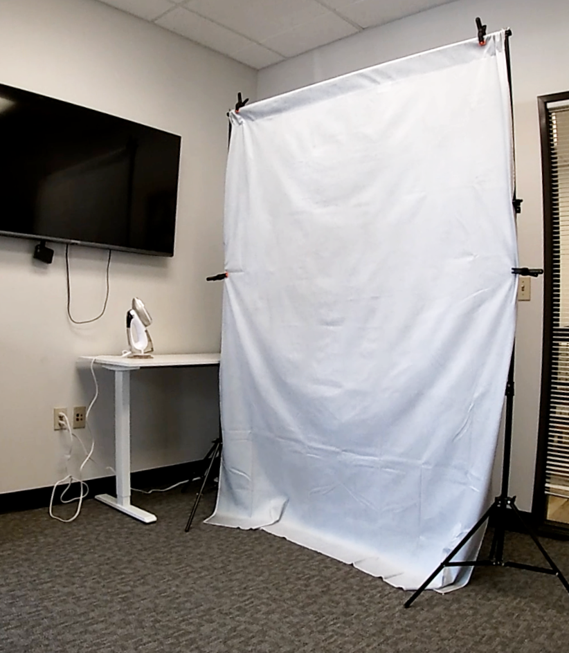 A white backdrop set on stands in an office, with a desk lamp on a nearby desk and a TV wall-mounted, creating a simple photo setup.