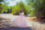 Quinceañera in a pink xv gown stands in a sandy, green, wooded area, wearing a tiara, smiling. Bright, sunny day, dress adorned with butterflies.