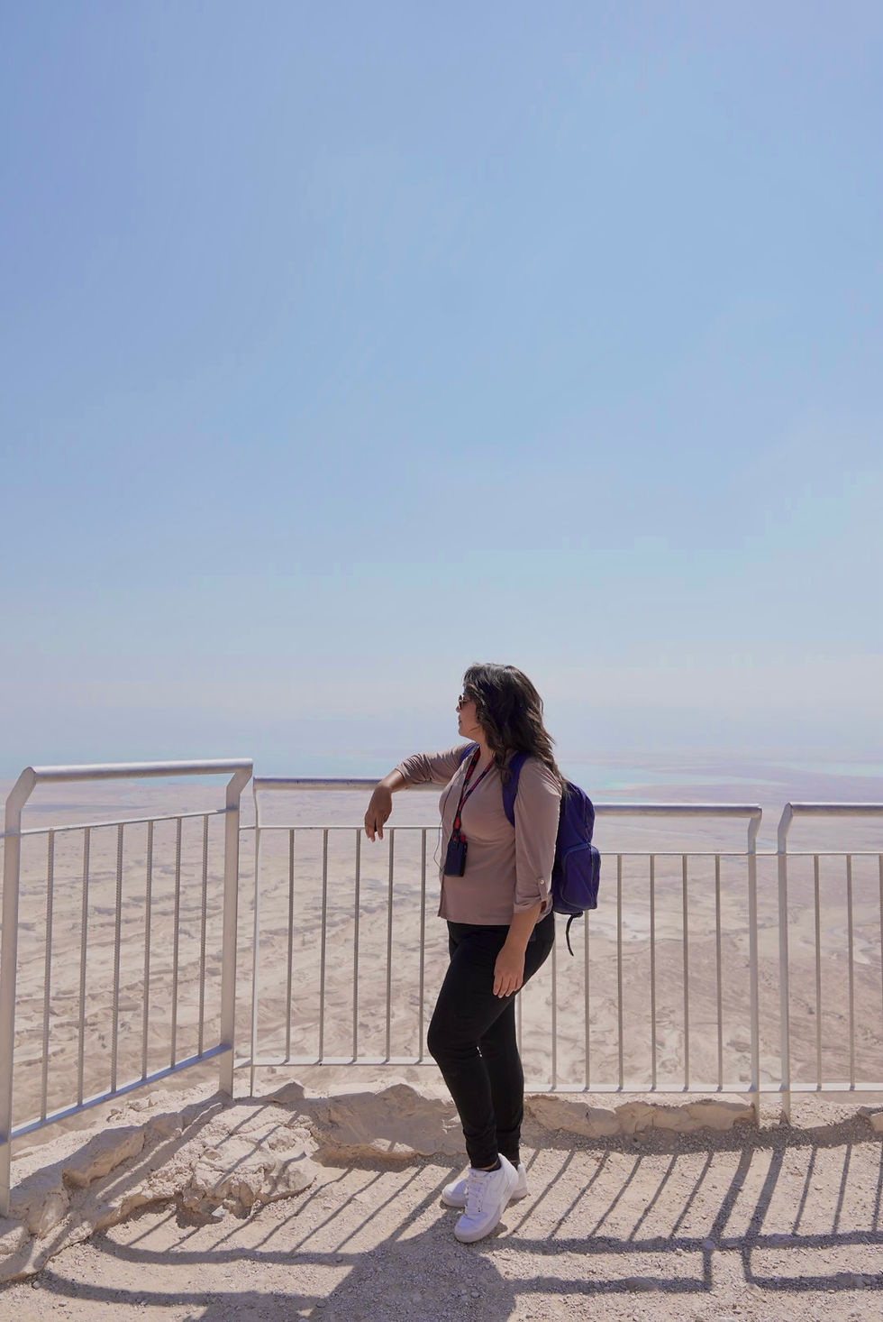 Woman in sunglasses leans on a railing at a hazy desert overlook, wearing a backpack and lanyard.