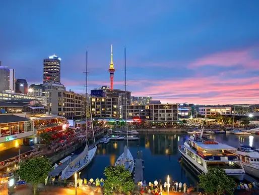 This image captures a vibrant dusk scene at Auckland’s Viaduct Harbour. The iconic Sky Tower dominates the skyline above luxury yachts, waterfront dining, and the city's glowing urban architecture.