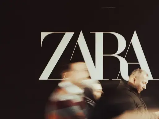 A dramatic, low-angle shot of the white Zara logo against a black background. Several blurred figures of people are captured in motion as they walk past the sign, creating a sense of a busy urban environment.