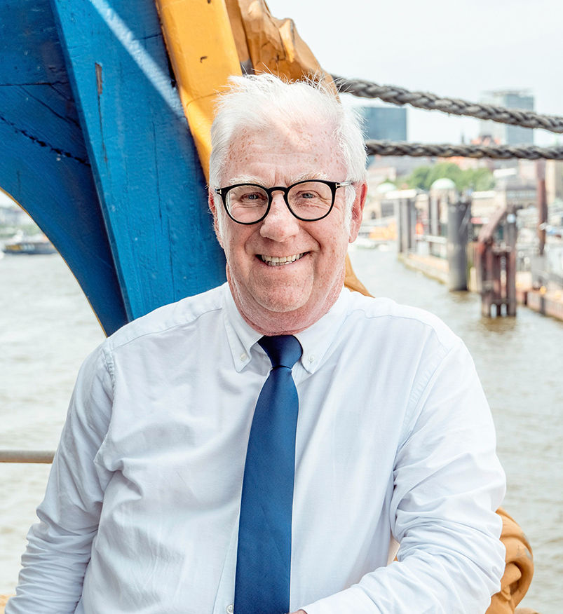 Medelålders man med glasögon och slips ler, bakgrunden är ett fartyg.