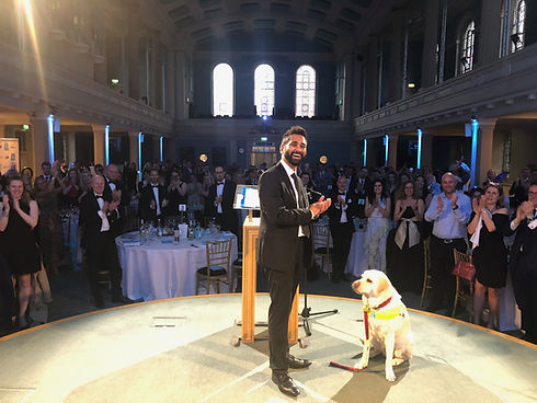 Dr Amit Patel stands on stage in a grand hall, dressed in a black suit and smiling as he claps in response to a standing ovation. Beside him, his guide dog sits calmly in harness, sharing the spotlight. The audience, also in formal attire, applauds warmly. Tall arched windows, illuminated columns, and a high ceiling create a dramatic, celebratory atmosphereāsuggesting an awards ceremony or gala event. The image captures a moment of recognition, pride, and partnership, highlighting both Amitās achievements and the quiet strength of his guide dog.