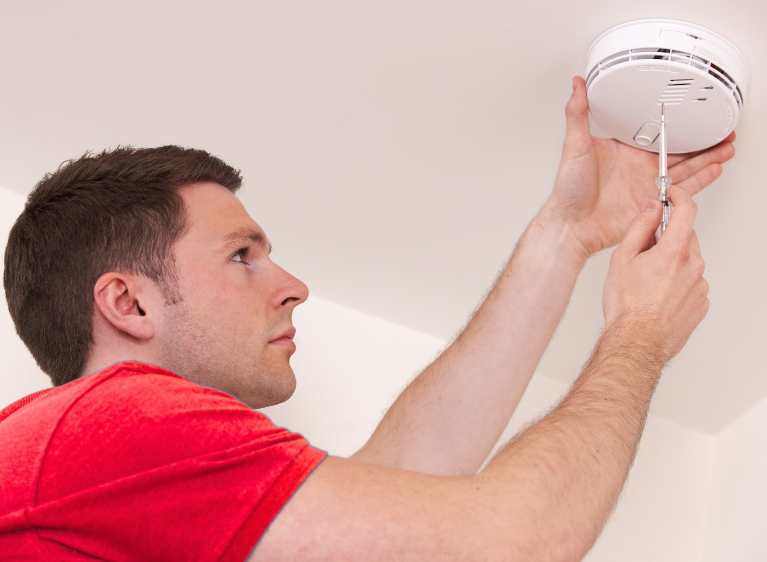 Man in a red shirt uses a screwdriver to install a smoke detector on a ceiling, focused indoors.