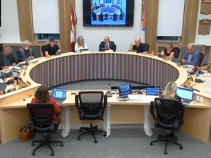 Council meeting in progress with people seated at a round table, laptops open. A screen displays images. The mood is focused and formal.
