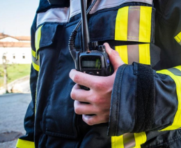 Firefighter in black and yellow gear holds a walkie-talkie. Outdoor background with clear sky. Focus on communication and readiness.