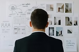 Man in a suit seen from behind studying a wall of website wireframes and interior photos in a bright office.