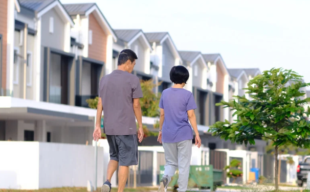 Two people in casual clothes walking on a suburban street lined with modern houses. A small tree is in the foreground.