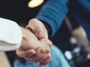 Two people in formal attire shake hands warmly, suggesting agreement or partnership. Blurred indoor background with soft lighting.