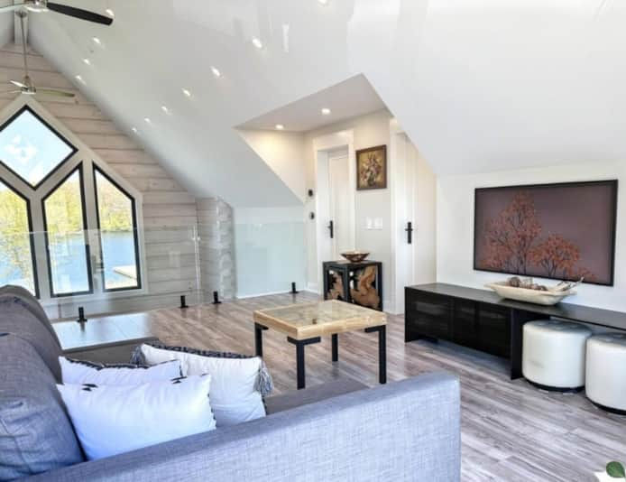Bright modern loft living room with gray sectional, wood coffee table, large windows, TV console, and sloped white ceiling.
