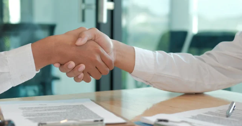 Two people in white shirts shake hands over a table with documents. The setting is an office with glass windows, creating a professional mood.