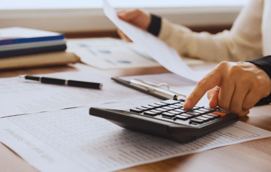 Hand using a black calculator on documents, one holding paper. Pen and clipboard nearby on a wooden desk. Office setting.