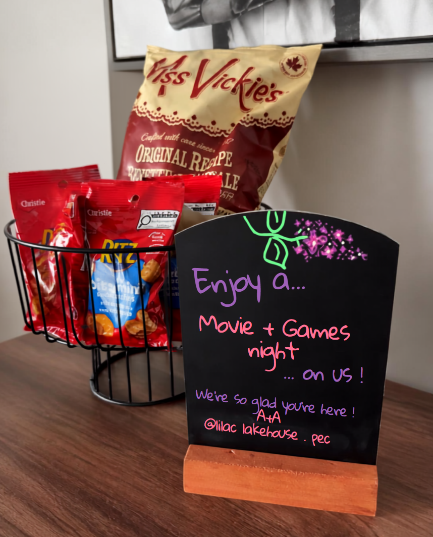 Snack basket with chips and cookies beside a black sign inviting guests to a Movie + Games night on a wooden table.