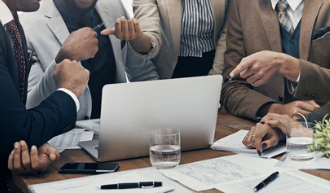 Business team in suits points at a laptop during a meeting, with papers, pens, and water glasses on the table.