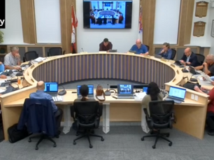 People seated around a horseshoe-shaped table in a meeting room. Screens show video conferencing. A logo reads "The County." Professional setting.