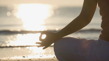 Person meditating cross-legged on a beach at sunset, hand in a calm mudra against glowing waves.