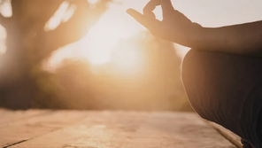 Person meditating outdoors at sunrise, sitting cross-legged with hand in a mudra pose. Warm sunlight and trees create a peaceful atmosphere.