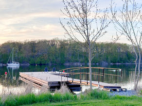 Quiet lakeside dock with two blue chairs, a moored sailboat, and trees at sunset under a pale sky