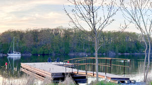 Quiet lakeside dock with two blue chairs, a moored sailboat, and trees at sunset under a pale sky