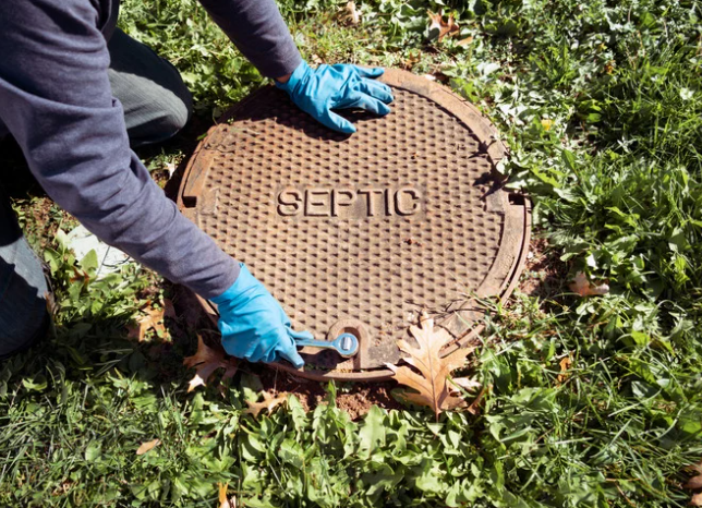 Person in blue gloves pries open a round septic tank cover labeled SEPTIC in a grassy yard