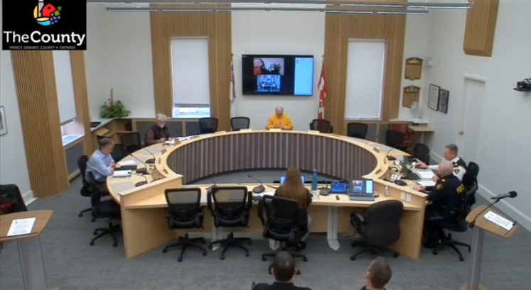 Council meeting in a circular room with six attendees seated. A screen shows a video call. Walls have plaques; Canadian flags visible.