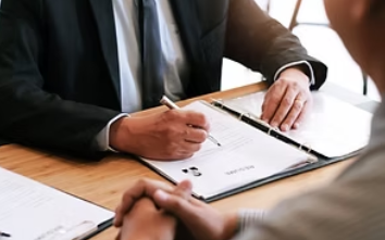 Business meeting with a man signing documents on a clipboard at a desk, while another person listens nearby.