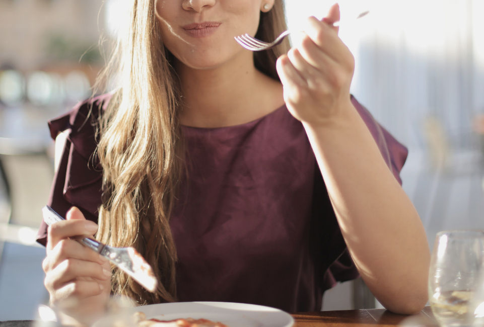 Woman in a purple shirt eats pasta at a brightly lit table, holding a fork and smiling. A green bottle and glass are in the foreground.