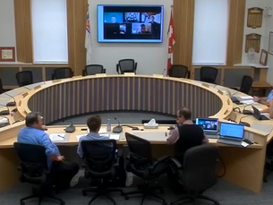 Five people sit around a circular conference table with laptops in a formal meeting room. A video call is displayed on a screen on the wall.