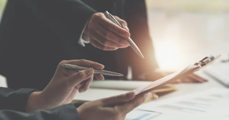 Hands holding pens over documents on a clipboard, with charts visible. Sunlight filters through an office setting, creating a focused mood.