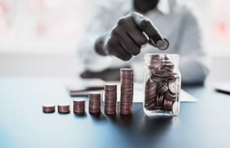 Hand placing a coin into a jar of coins beside rising stacks on a table, with a blurred person in the background.