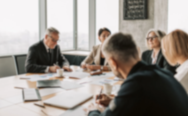 Blurred image of a business meeting with five people discussing around a conference table. Large windows and cityscape visible in the background.