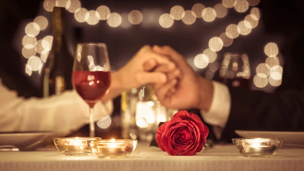 Romantic dinner setting with a couple holding hands, red wine glasses, red rose, and candles on the table. Warm, intimate ambiance.