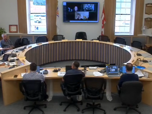 Seven people sit in a circular meeting room with laptops; a virtual meeting is on a screen. The room has large windows and flags.