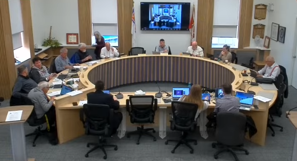 Meeting with 12 people in a round conference room. Laptops open; a screen displays a video call. Neutral tones and professional setting.
