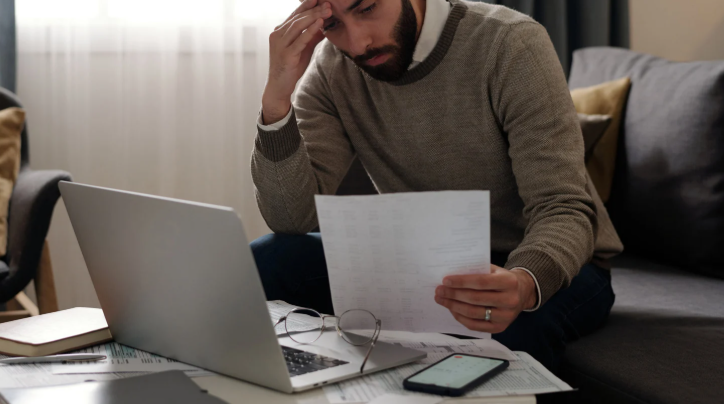 Man sitting on sofa, looking stressed while holding papers, laptop open, phone nearby. Neutral-toned room with soft lighting.