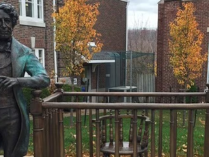 Bronze statue of a standing man beside a chair on a terrace. Brick building and autumn trees in the background. Overcast sky.