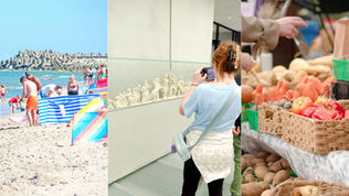 Beachgoers by sea wall, a woman photographs a sculpture display, and a market stall shows baskets of produce.