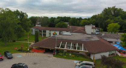 Aerial view of a large brown-roofed building surrounded by trees and parked cars. Overcast sky and grassy areas create a calm atmosphere.