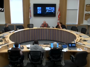 Circular conference room with eight people seated at a round table. A screen displays images. Flags and logo of The County are visible.