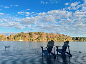 Two black Adirondack chairs on a dock by a calm lake, facing autumn trees under a blue cloudy sky; SnapEdit watermark.