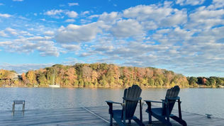 Two black Adirondack chairs on a dock by a calm lake, facing autumn trees under a blue cloudy sky; SnapEdit watermark.
