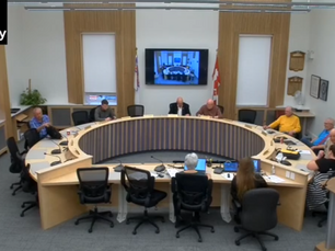 Council meeting in a circular room with six attendees seated. A screen shows a video call. Walls have plaques; Canadian flags visible.