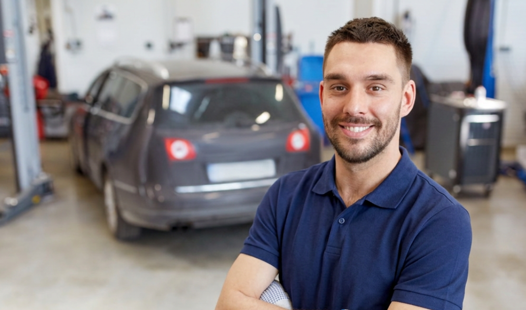 Smiling mechanic in a blue polo and gloves stands in an auto shop with a car on a lift behind him.