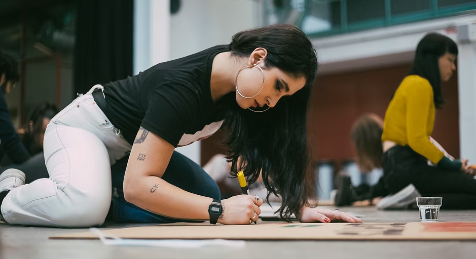 A woman in white jeans and a black shirt writes on cardboard on the floor. Other people are doing the same in a well-lit room.
