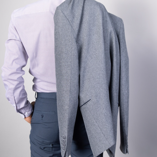 Man seen from behind in dress pants and pale shirt, a gray blazer draped over one shoulder against a plain studio background