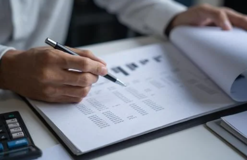 Person reviewing financial documents with a pen, calculator nearby on a desk. The setting is an office, with a focused, analytical mood.